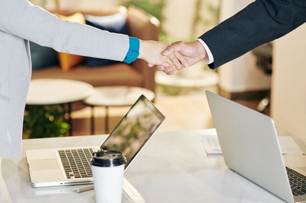 image of two people in business attire shaking hands over laptops on a table. They may be potential directors discussing how many directors a limited company can have and whether this will benefit their company.