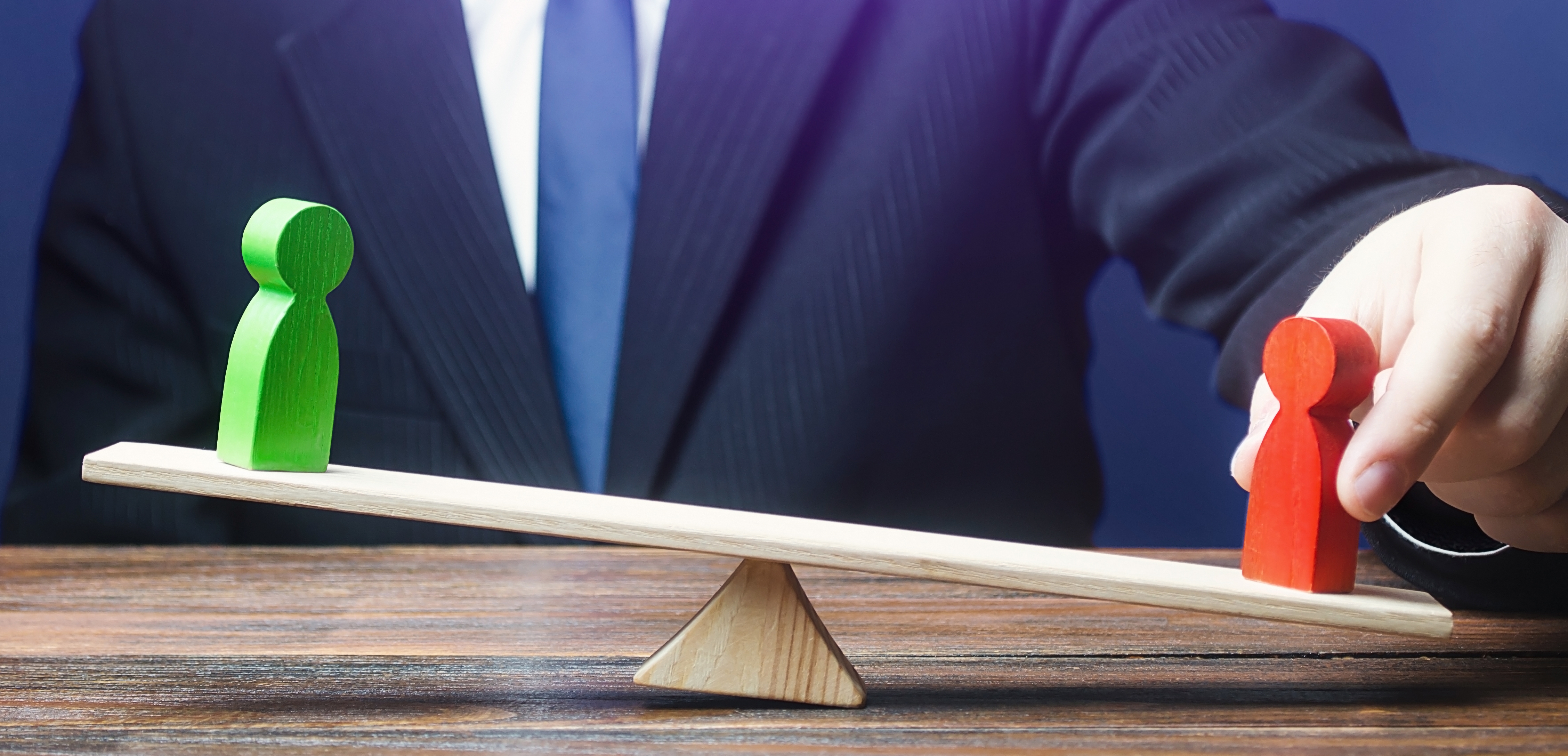 Two wooden figures, left (green) and right (red) on a wood plank which is weighted to the right on a wood triangle block. There is a person in a suit holding the red figure. This insinuates the answer to ‘do all shareholders get equal dividends’