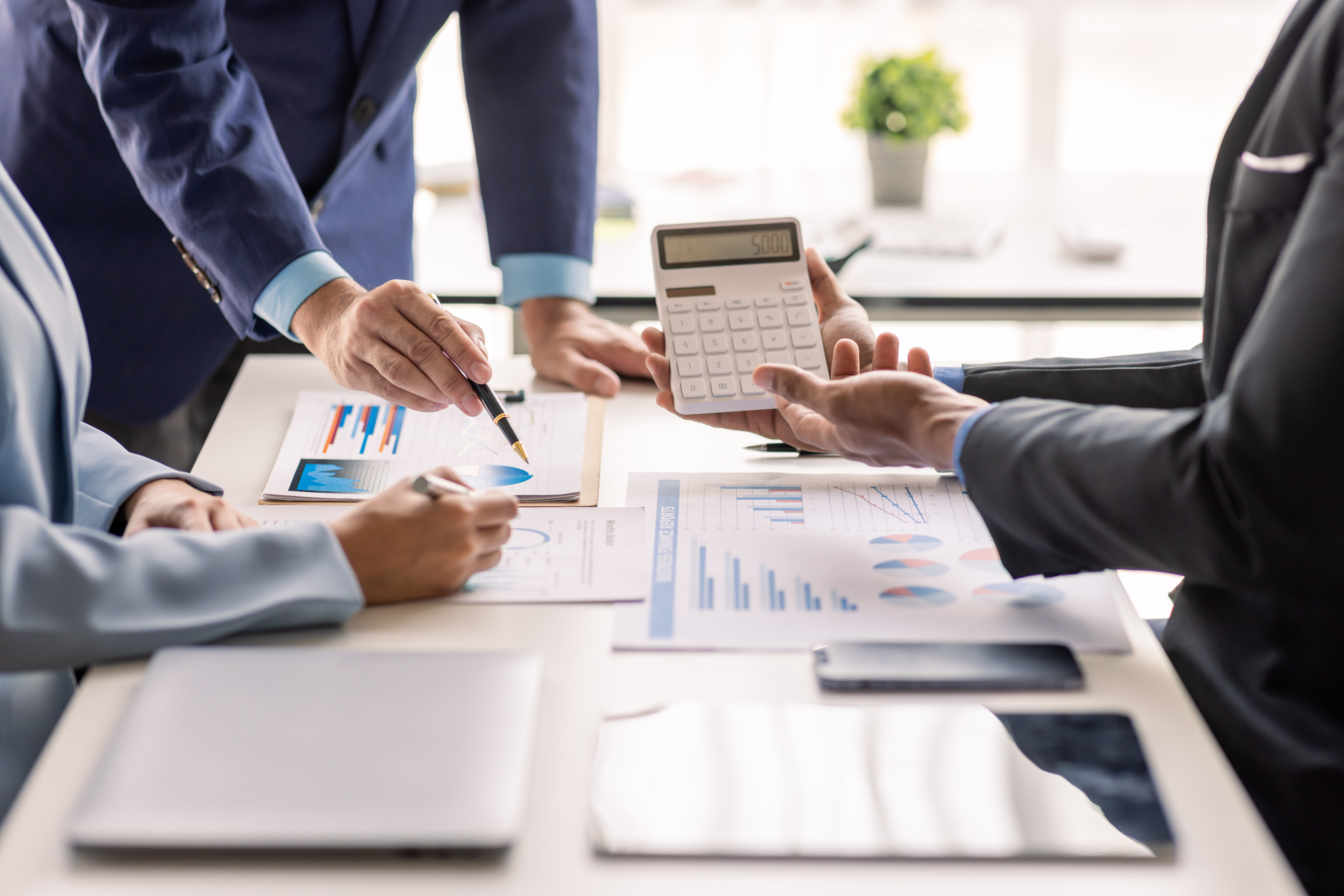 Three Limited company directors stood over a table, with documents, a calculator, laptop and pens. They are discussing tax-deductible expenses in their limited company.