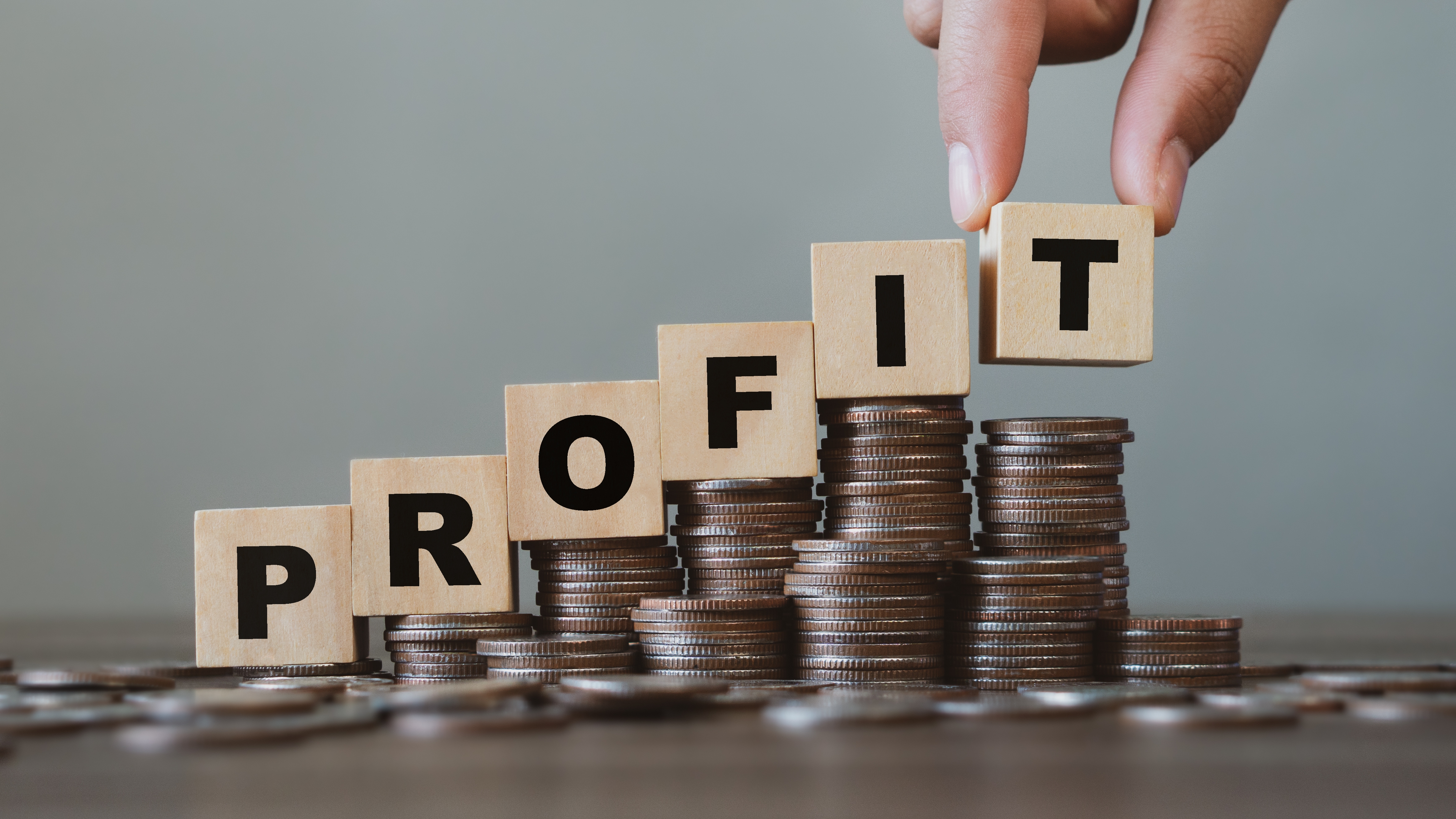 A wooden table with coins scattered and stacked, on top of the stacks of coins are wooden blocks spelling out ‘PROFIT’ in an ascending order. The ‘T’ in profit is being placed down by two fingers.